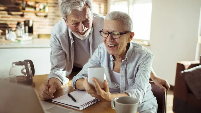 Shot of two seniors sitting at table looking at a phone with a laptop and notebook in front of them