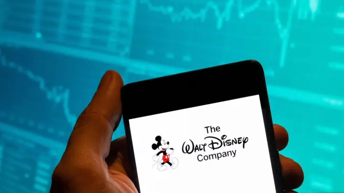 In this photo illustration, the American multinational mass media and entertainment The Walt Disney Company or also called Disney logo is seen displayed on a smartphone with an economic stock exchange index graph in the background.