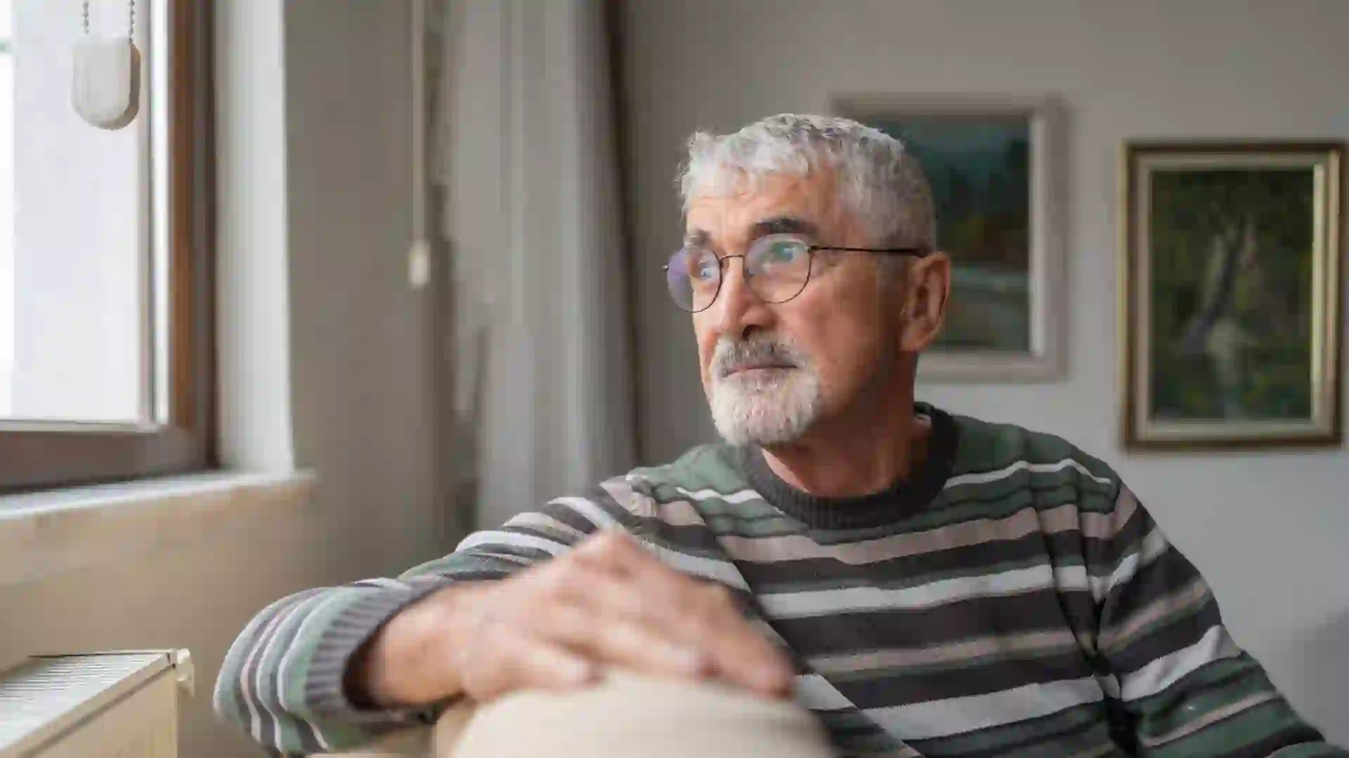 Senior Man Sitting On Sofa By Window And Looking Outside Through Window At Home.