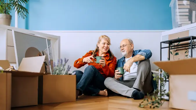 A joyful senior couple sits together on the floor, surrounded by cardboard boxes and personal belongings.