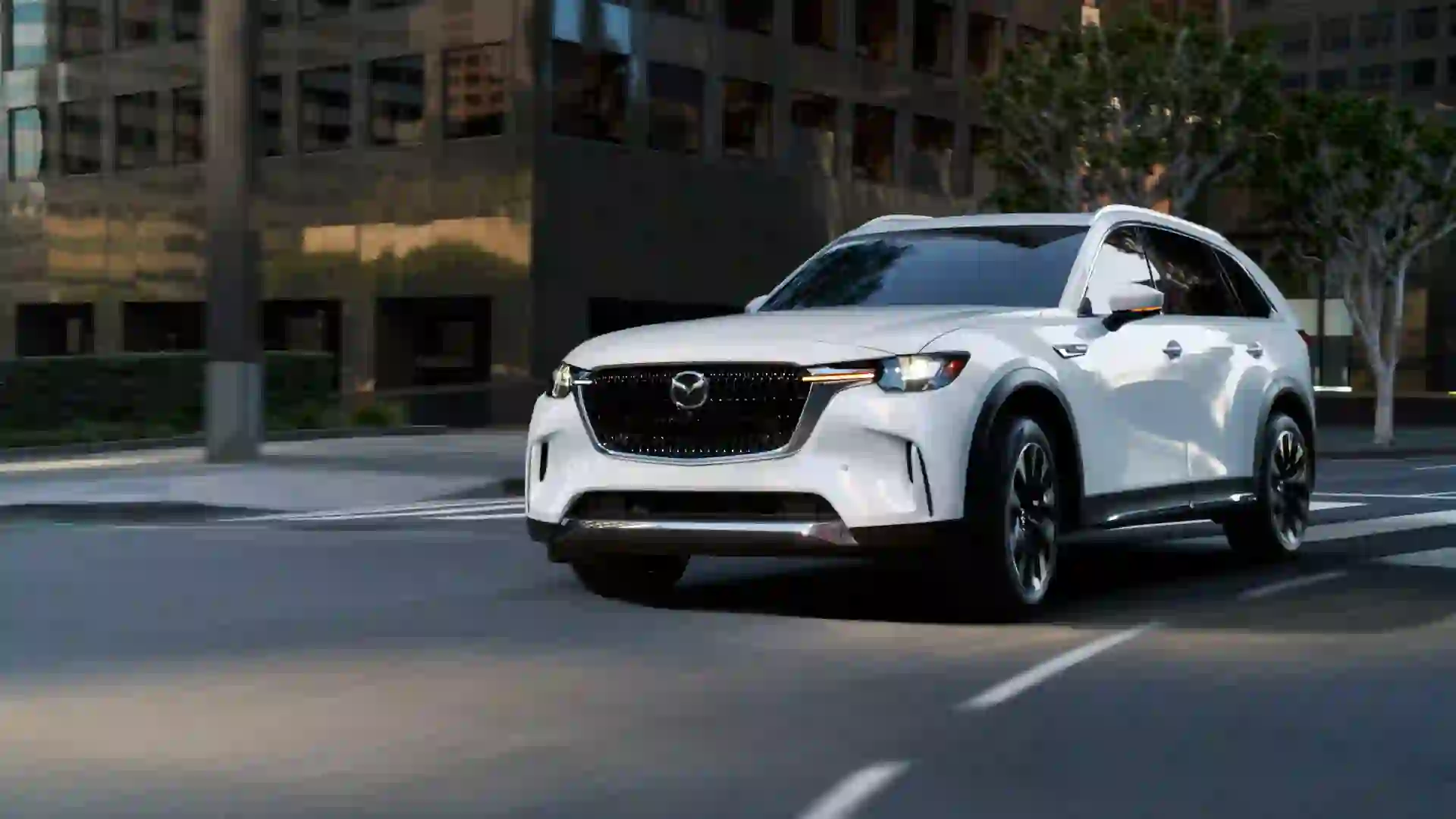 A white 2025 Mazda CX-90 driving down a city street for promotional photo.