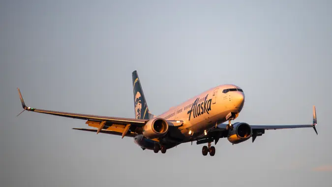 A close-up of an Alaska Airlines Boeing 737-800 as it approaches for landing at the John Wayne Airport in Santa Ana, California, bathed in golden hour sunlight.