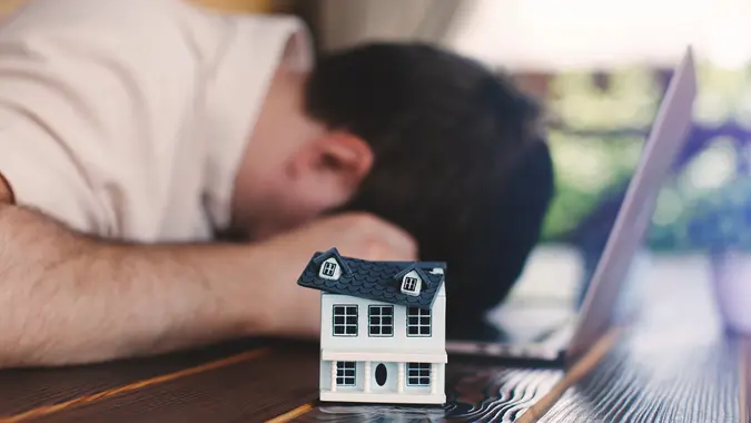 Upset man on a table with a laptop and a model of a house whose roof has broken down. Home renovation concept stock photo
