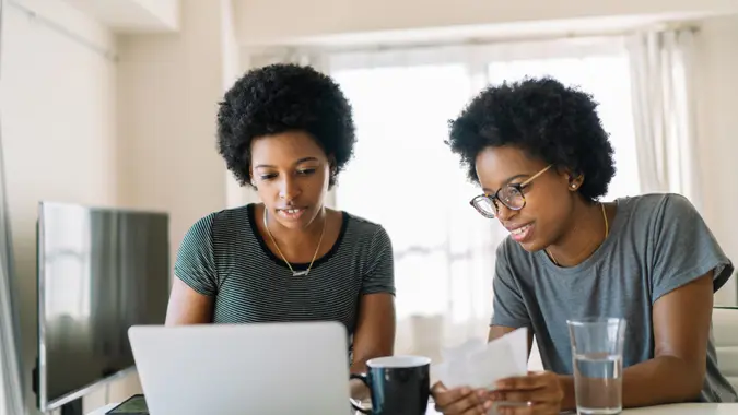 Two black female twin sisters are doing home finance on a laptop in the living room at home.