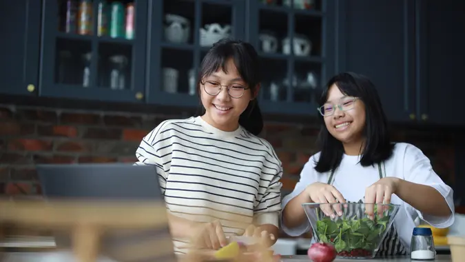 A cheerful young Asian sister is using a laptop computer in the kitchen to choose what to cook for lunch, while also mixing salad in a bowl and watching a culinary video blog.
