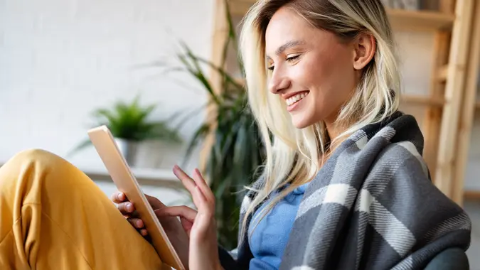 Happy young woman with blanket over her shoulders happily using a tablet computer in her loft apartment