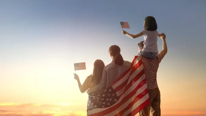 A family looking toward a sunset, holding an American flag across their backs and smaller flags in their hands.