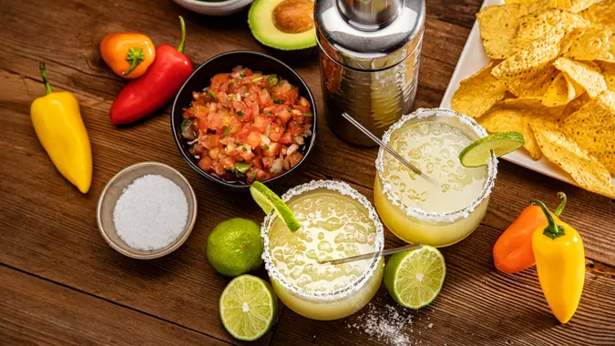 This is a photograph of two modern margarita glasses with a rim of salt surrounded by fresh cut limes and chips and salsa on a retro wood background photographed from a high camera angle.