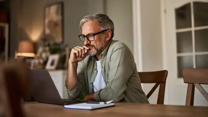 A focused mature man at home sitting at his wooden dining room table using his laptop to go over finances
