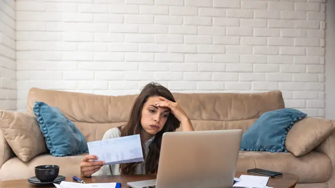 Young woman sitting on the floor in front of a couch, looking at a laptop and papers with one hand on her forehead and a stressed expression.