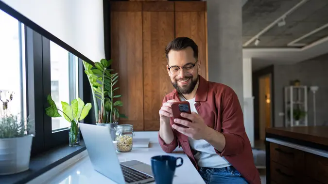 Photo of a Man with glasses using his mobile phone while siting and working on his laptop in his home.