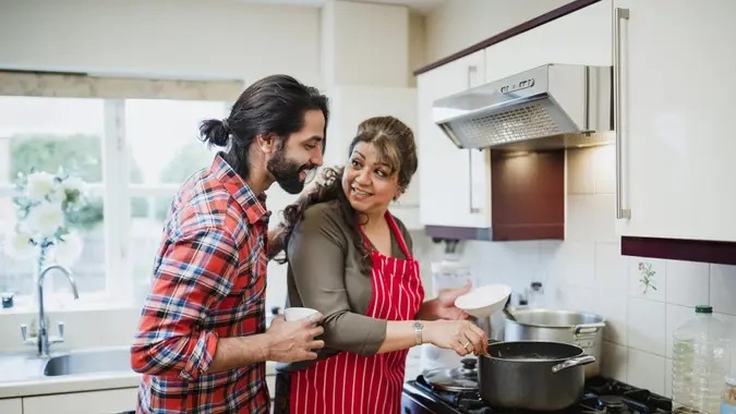 Mature woman cooking in her kitchen while her adult son looks over her shoulder.