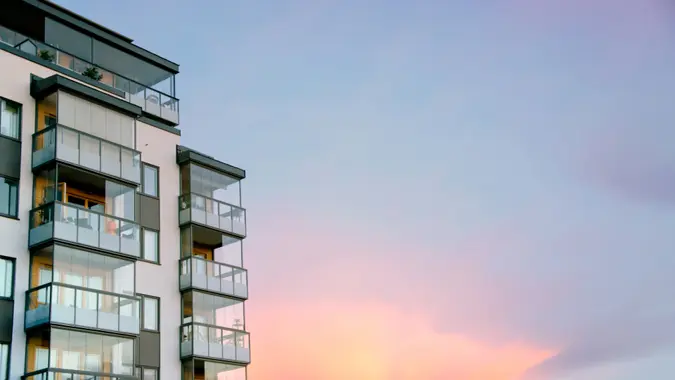 Behind a modern condo building, the sun is setting, painting the clouds in vibrant colors.