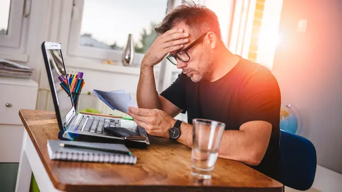 Man sitting at table with laptop in front of him is reading letter and feeling worried over finances, bills, budgeting and more.