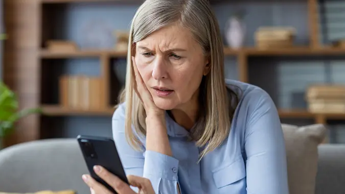 Worried senior woman sitting on a sofa while looking at a smartphone with a confused or worried expression.
