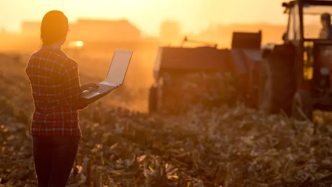 Young woman with laptop standing on field in sunset and looking at tractor baling.