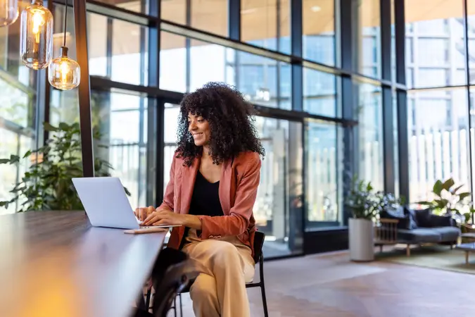 Picture of a Young woman sitting at a desk using a laptop for her finances in a window lined office