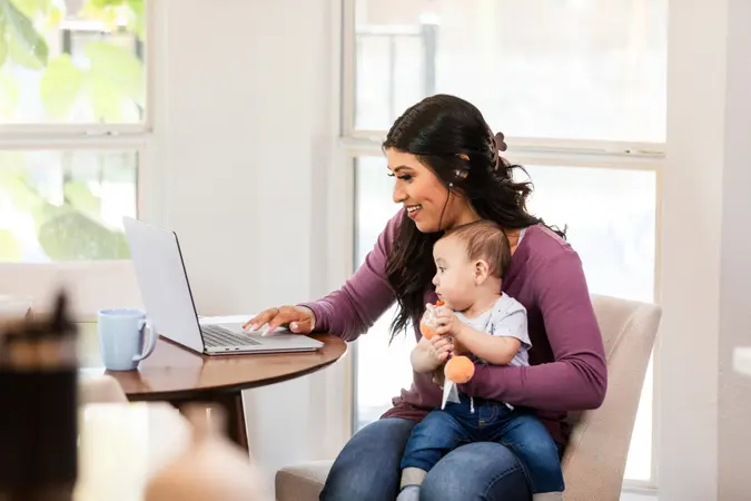 A cheerful businesswoman smiles as she uses a laptop while holding her infant son.