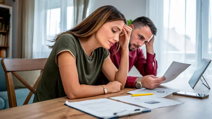 A young couple with worried expressions sits at a dining table, reviewing financial paperwork with a tablet and phone.