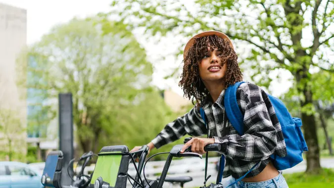 Cheerful young latin student with backpack and camera is ready to ride a bike sharing in the city.