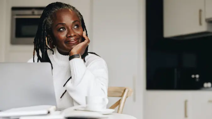 Mature businesswoman looking away thoughtfully while working on a laptop at home.