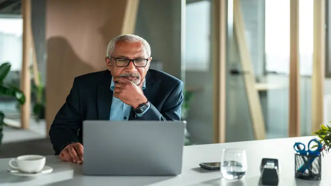 A senior mixed-race businessman reflects deeply as he works on his laptop, dressed professionally in a business environment.