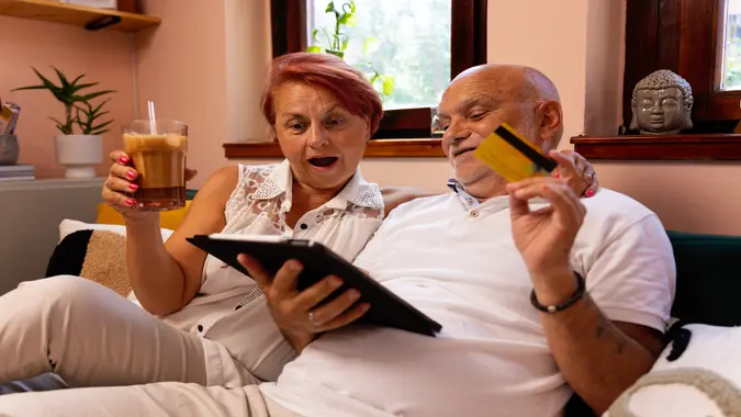 A lovely senior couple sits together in their cozy living room, using a tablet for online shopping.