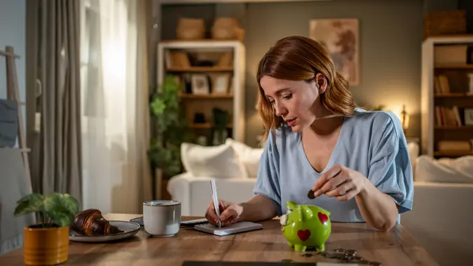 Young woman carefully managing her home finances, putting coins in a piggy bank and taking notes on expenses and savings in her living room.