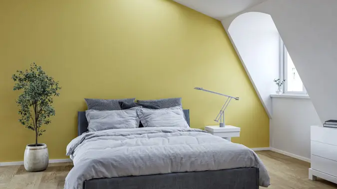 Modern minimalist bedroom in the attic with light wooden flooring, yellow wall background with copy space, neutral bedding, and a cozy atmosphere.