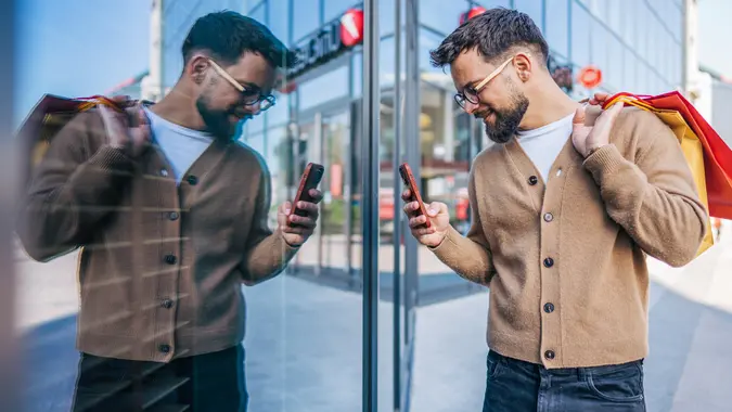 Stylish man using smartphone while carrying shopping bags in front of modern building stock photo