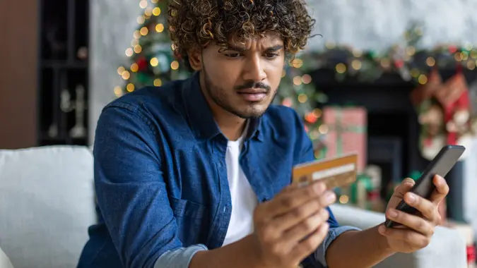 Cheated and upset Christmas man in living room near decorated Christmas tree, rejected and wrong money transfer, hispanic man holding bank credit card and phone stock photo