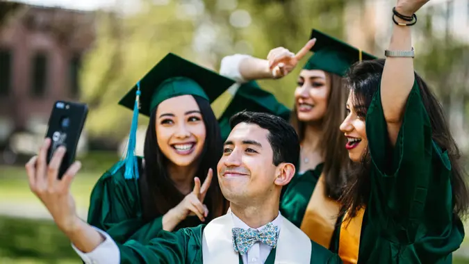A multiracial group of male and female college graduates pose together and take a selfie photograph with a smart phone outside on graduation day.