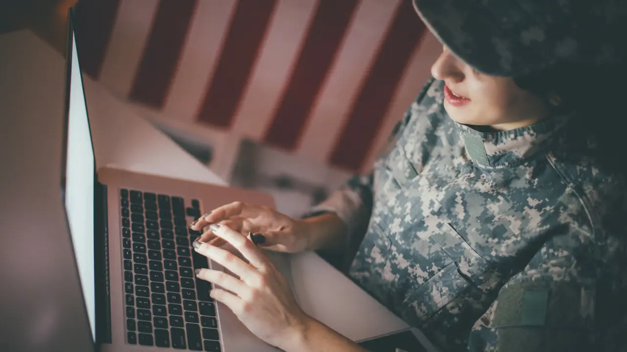 Woman in military uniform types on a laptop with an American flag in the background, working in a dimly lit room.
