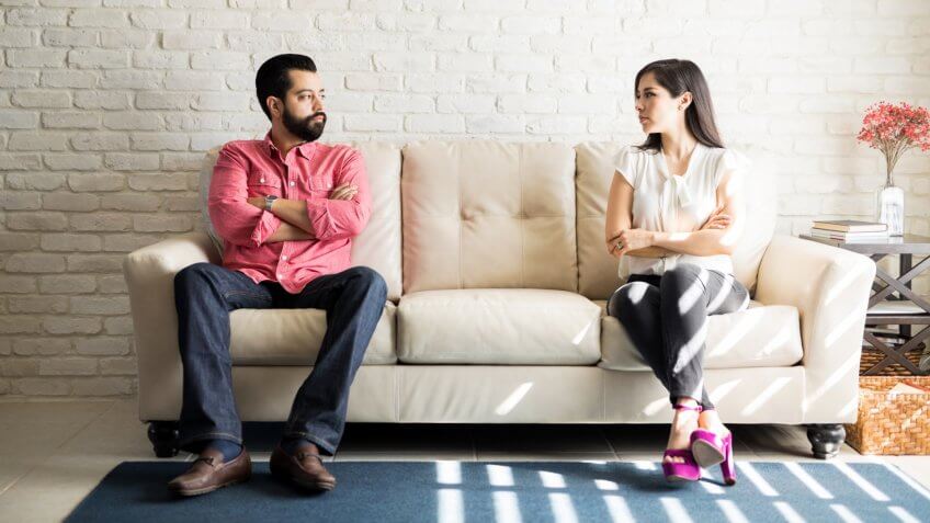 Husband and wife sitting on sofa with their arms crossed and looking at each other after a fight in living room.