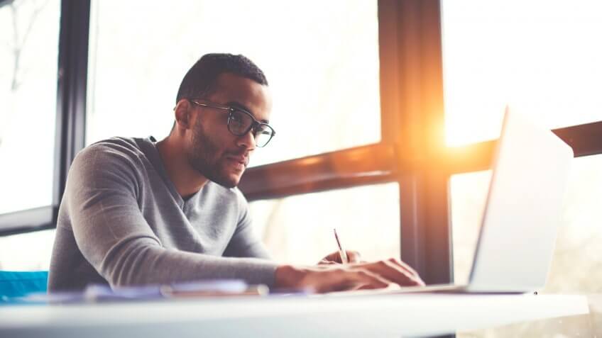 Concentrated dark skinned copywriter in eyewear typing text of publication for web site working on freelance, serious afro american student searching information for homework project in network.