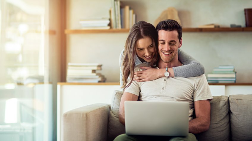 Shot of a young woman hugging her husband while he uses a laptop on the sofa at home.