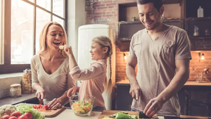 Cute little girl and her beautiful parents are cutting vegetables and smiling while cooking in kitchen at home - Image.