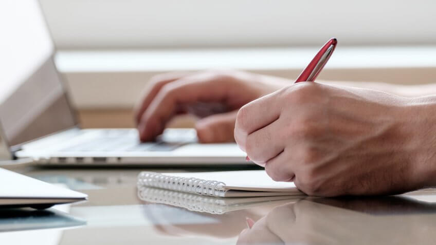 Close up of male hands with laptop computer, notebook and pen taking notes in business office.
