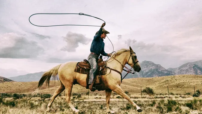 Young cowboy with lasso riding quarter horse on the open western range with mountains in the background.
