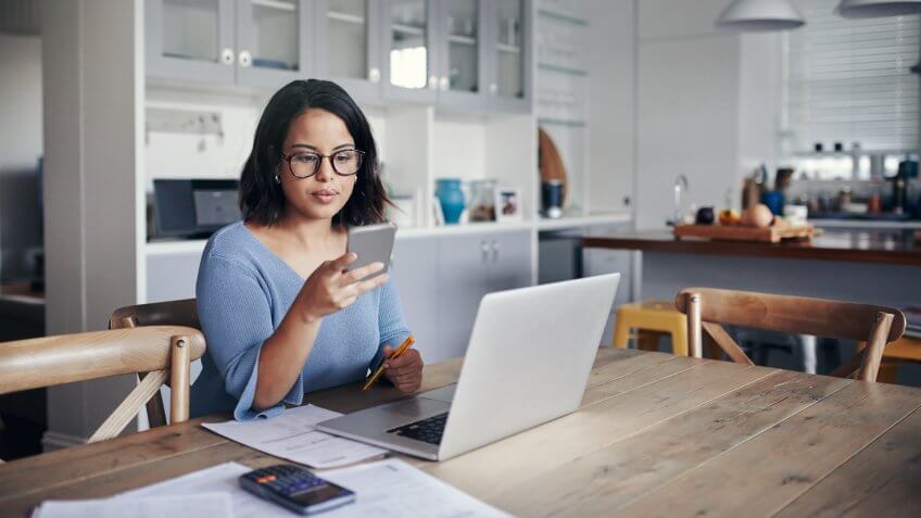Shot of a young woman using a mobile phone and laptop while working from home.