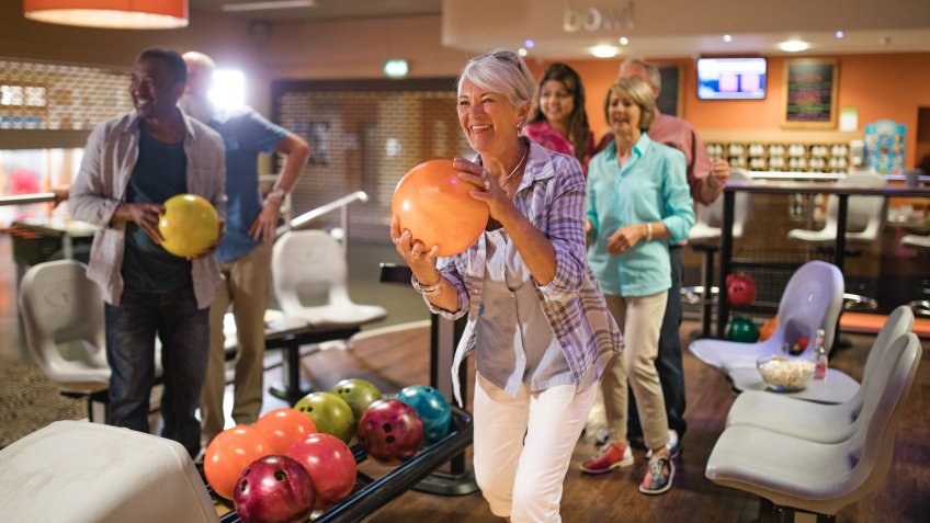 Group of seniors at a bowling alley, five senior friends can be seen in the background whilst a woman takes her turn.