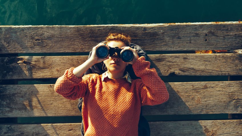 Young Girl Lies On A Pier Near The Sea And Looks Through Binoculars.