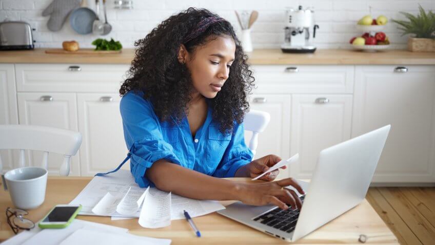 Candid shot of seriousn concentrated young African female using modern technology while paying for gas and electricity online, hoding receipt and typing on laptop.