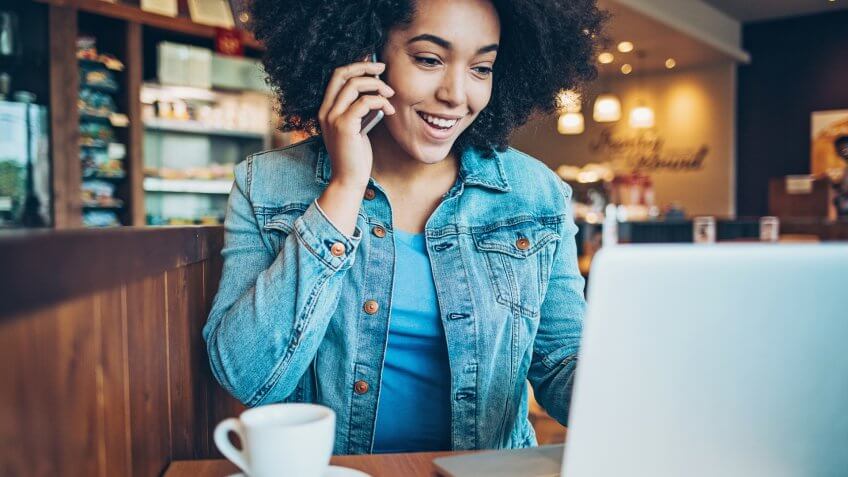 Smiling young African ethnicity woman using smart phone and laptop in cafe.